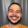 Foto exibindo o rosto do Alvaro Vasconcelos. Ele está vestindo uma camiseta laranja. O fundo da imagem é de cor cinza com uma textura sutil, possivelmente uma parede ou cortina.
